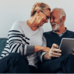 An elderly couple sits closely on a teal sofa, smiling and looking at a tablet together. The woman, wearing glasses and a striped sweater, leans towards the man, who is wearing a dark vest over a shirt. Both appear happy and engaged, perhaps perusing the services options of their trusted advisory firm.