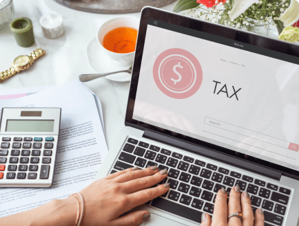 Someone is using a laptop with a tax website on the screen. Beside the laptop, there is a calculator and some papers. A cup of tea, a pair of glasses, and a flower arrangement are also visible on the table, creating a work-from-home setting.