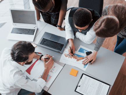 Four people are gathered around a table working collaboratively. They have laptops, documents, and charts in front of them. Two individuals are pointing at a graph, while the other two observe. The atmosphere appears to be focused and engaged.