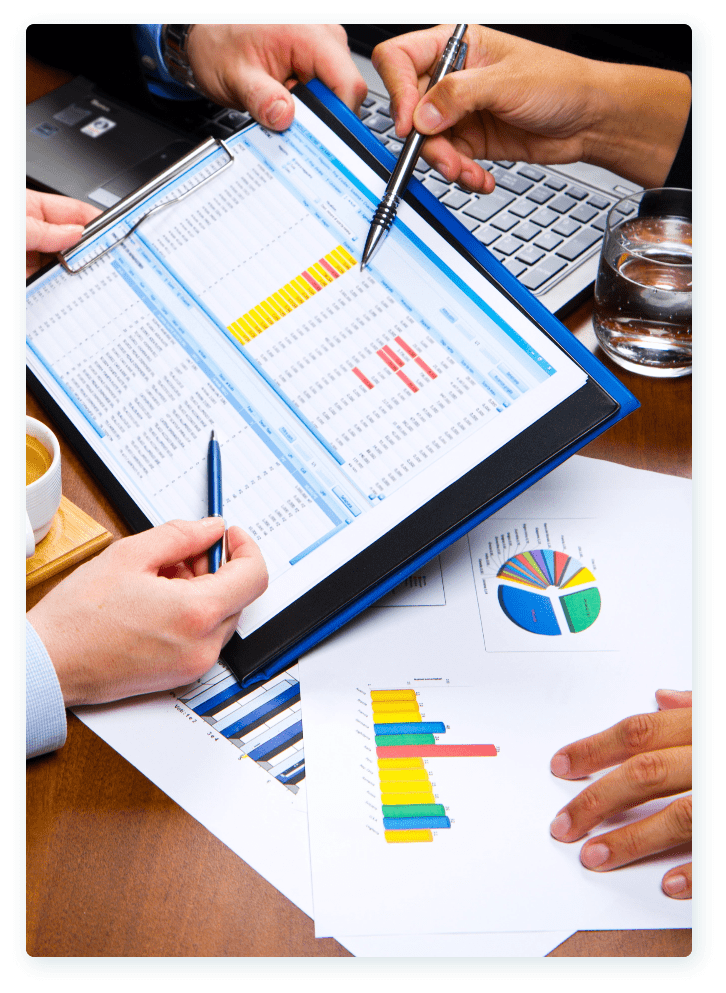 Three people are seated at a table engaged in a discussion. One person is holding a clipboard with a spreadsheet, while another points at it with a pen. Nearby, there's a laptop, documents with graphs, a keyboard, a glass of water, and a cup of coffee.