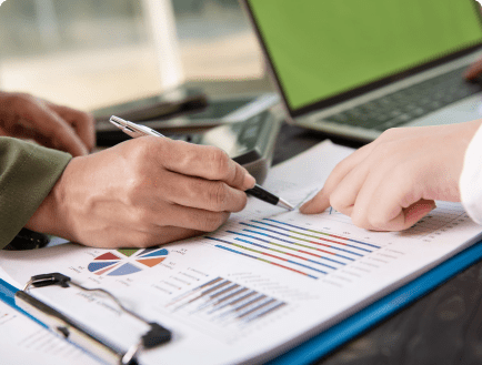 Close-up of two people's hands pointing at charts and graphs on a paper. One hand holds a pen while the other points at a specific section on the paper. A laptop and other office supplies are visible in the background.