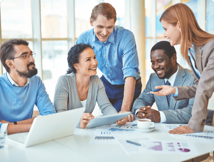 A group of five people are gathered around a table in a bright office space. They are smiling and engaged in a discussion, with documents and a laptop in front of them. One person is holding a tablet and another is presenting, creating a collaborative atmosphere.