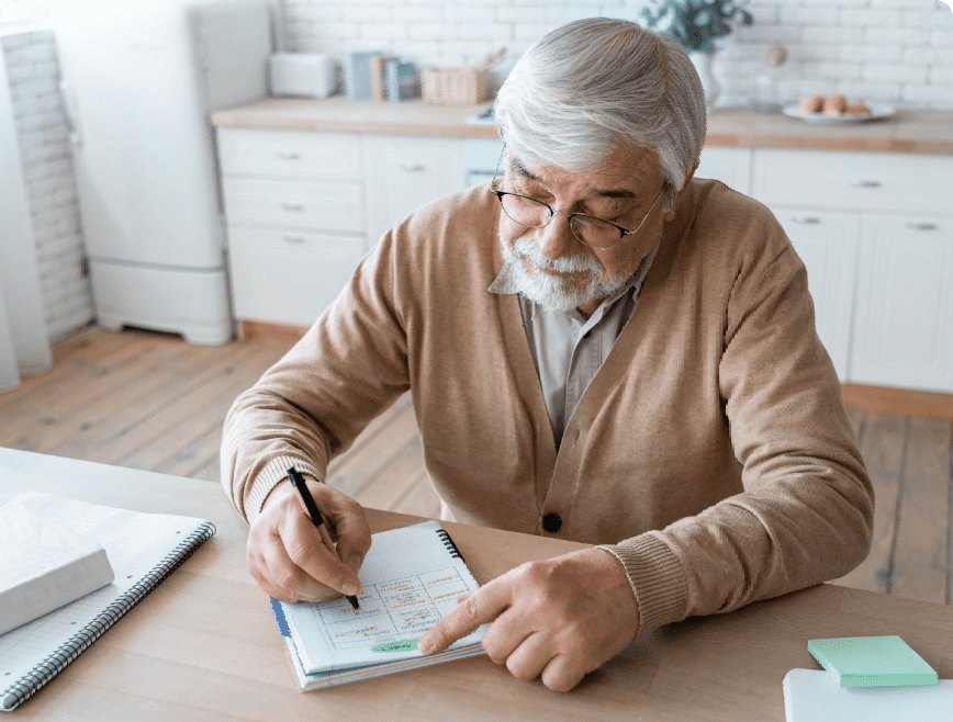 An elderly man with grey hair and glasses is sitting at a kitchen table, writing in a notebook. He is wearing a beige cardigan over a collared shirt. His other hand points at the notebook, which contains written notes, as he appears focused on his task.