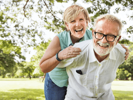 A joyful older couple, both wearing glasses, smile and pose playfully in a sunny park. The man is piggybacking the woman, and they are both laughing. Green trees and a grassy field are visible in the background.