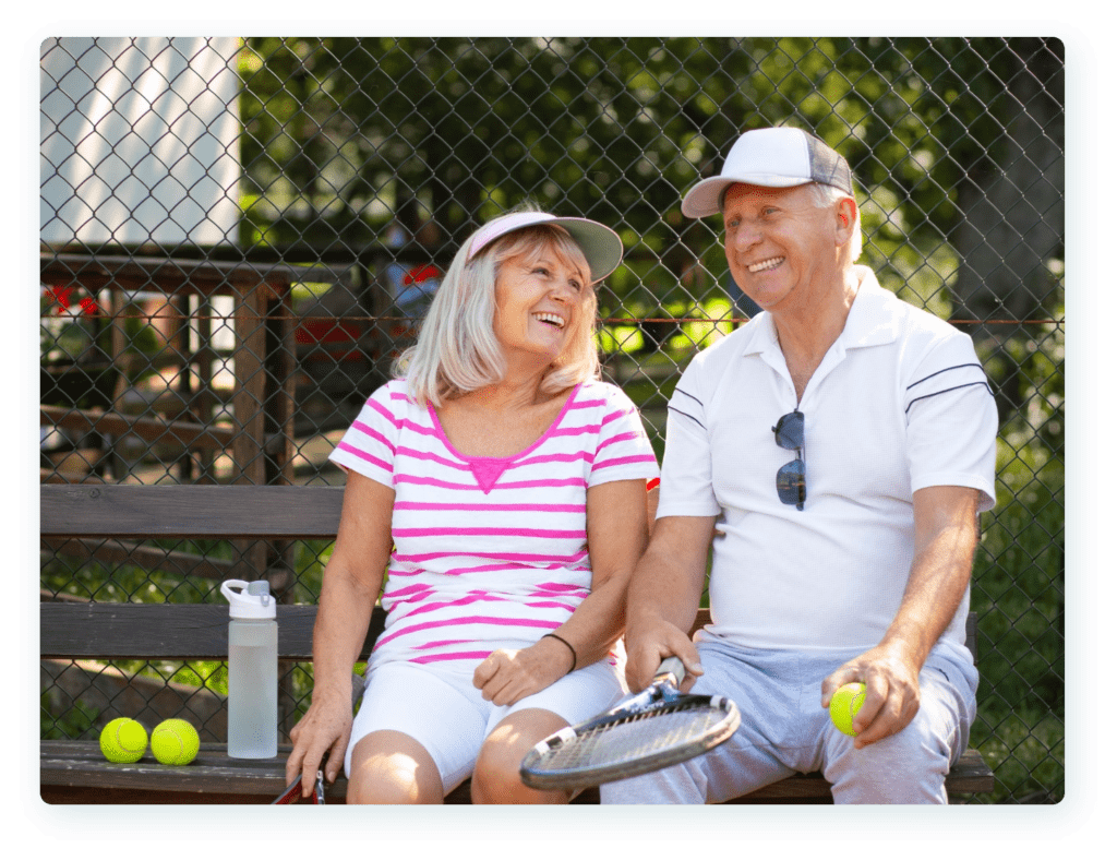 An elderly couple is sitting on a bench by a tennis court, smiling and laughing. The woman is wearing a pink-striped shirt and cap, while the man is wearing a white shirt, cap, and has sunglasses hanging from his collar. Tennis balls and a racket are near them.