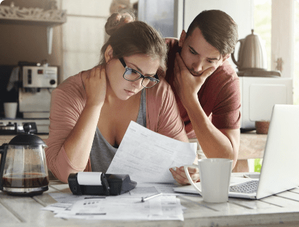 A concerned man and woman review paperwork at a kitchen table. The table is cluttered with a coffee pot, calculator, cup, documents, and a laptop, indicating they are working on financial or administrative tasks. Both appear thoughtful and focused.