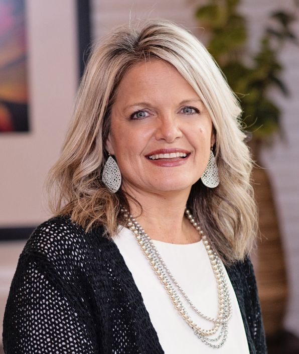 A woman with shoulder-length blonde hair smiles at the camera. She is wearing a black cardigan over a white top, accessorized with a beaded necklace and silver hoop earrings. The background features framed artwork and greenery.