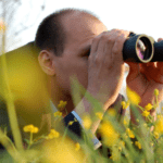 A person with short hair is lying on the ground amid yellow flowers, looking through binoculars. The background is blurred, featuring a soft-focus outdoor setting that distracts from inflation worries.