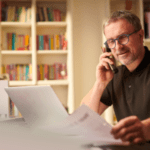 A man with glasses and a beard sits in front of a laptop in a home office with bookshelves in the background. He is smiling while talking on the phone and holding a sheet of paper. The ambiance suggests a relaxed yet productive work-from-home setting.