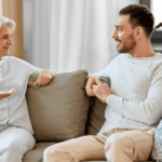 A senior woman and a younger man are sitting on a beige sofa in a cozy living room, likely discussing the joys and challenges of caring for aging parents. They are engaged in a friendly conversation, both smiling. The room features a white curtain, wooden shelves with various items, and a large green potted plant.