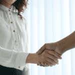 A person in a white long-sleeve shirt and black pants is shaking hands with another person whose arm is visible. The background is bright and softly lit, suggesting a professional or formal setting.