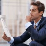 A man in a suit is sitting outdoors, holding a folded newspaper in one hand and resting his chin on his other. He appears to be deep in thought, perhaps pondering the complex taxation of stock options, with a blurred building facade in the background.