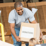 A man is smiling and holding a cardboard box labeled "Donations" while working in a warehouse. Numerous more boxes are stacked in the background. Another person in the foreground is partially visible, also handling boxes for this nonprofit organization, showcasing their investment in community support.