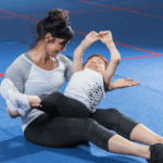 A woman is sitting on the gym floor, holding a laughing child who is lying on her lap with arms raised, on a blue mat with red lines in the background. Both are wearing athletic clothing and appear to be having fun together, capturing a happy moment that underscores the importance of special needs planning.