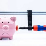 A pink piggy bank with a sad facial expression is being squeezed tightly by a large clamp. The clamp has a red and blue handle, suggesting financial pressure or constraints reminiscent of the recession in 2022. The background is plain white.