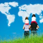 A family of four stands on a grassy hilltop, with two adults carrying two children on their shoulders. They face a clear blue sky where clouds form the shapes of the world map, illustrating the continents and reflecting on global events like economic pain in Russia.