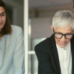 An older woman with short gray hair, a female investor in a black suit, is speaking while two younger colleagues, a woman in a light blue shirt and a man with a beard, listen attentively. They appear to be in a professional setting.