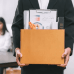 A person in a suit is holding a cardboard box filled with office supplies, including folders, a pen holder, and a few other items. In the background, another person is sitting at a desk. The scene suggests the individual may be exiting corporate life.