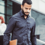 A man in a dark, patterned shirt looks down while walking outside a building, holding a laptop or briefcase. In the background, a woman with red hair is talking on her phone near the building's entrance, possibly discussing succession planning. The scene appears business-oriented.