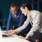 A man and a woman are working together at a desk, both looking at a laptop screen. The man, wearing glasses and a blue suit, is standing with his hands on the desk. The woman, in a beige suit, is leaning over, pointing at the screen and smiling as they discuss S corporation advantages.