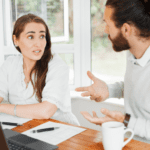 A woman and a man are having a discussion at a table. The woman gestures with her right hand raised and appears to be explaining something about financial planning. The man also gestures with his hands. They have papers and a laptop in front of them. The setting looks like an office.