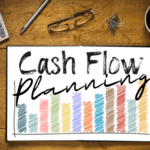 A wooden desk holds a calculator, glasses, paper clips, a cup of coffee, and a paper titled "Cash Flow Plan" with colorful bar graphs beneath the text.