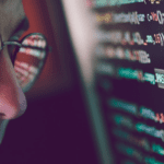 A person wearing glasses examines a computer screen filled with lines of code. The code is reflected in the glasses, highlighting the details of the text, including the word "PASSWORD" visible on the screen. The focus is on the tech-related activity, emphasizing cyber security measures.