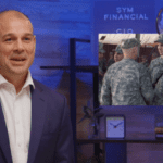 A man in a suit stands to the left, smiling. Behind him, a screen displays a photo of soldiers in military attire standing together, possibly with their platoon leader. The background features a blue wall with "SYM FINANCIAL" and "CIO" written on it, along with a clock and decorative items on a shelf.