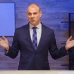 A man in a navy suit and blue tie stands behind a desk, gesturing with both hands. The background features a wooden wall, a whiteboard on the left, and a shelving unit on the right with books and decor items. The scene appears to be in an office or studio setting, likely discussing the volatile market conditions.