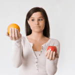 A woman with long brown hair, wearing a white long-sleeve shirt, is holding an orange in her left hand and a red apple in her right hand. She appears to be contemplating the two fruits. The background is plain white.