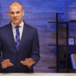 A man in a navy suit and blue checkered tie is speaking and gesturing with his hands. He stands behind a desk with a sleek, minimalist background, including shelves with books, a clock, and a plant. A sign reads "SYM Financial CIO Corner" in the top right.