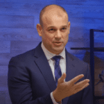 A bald man in a blue suit and tie is speaking animatedly, gesturing with his right hand. He is standing in front of a wooden textured wall with a blue tone, discussing artificial intelligence. A black shelf can be seen in the background.