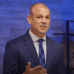 A man in a blue suit with a white shirt and blue checkered tie is speaking. Gesturing with his right hand, he stands against a wood-paneled wall accented by blue lighting and a black shelf. Discussing artificial intelligence, his words resonate within the modern setting.