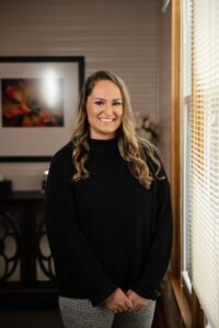 A woman with long, wavy hair and wearing a black sweater and patterned pants stands smiling near a window with blinds. The background includes a framed abstract artwork on a wall with wood paneling and a piece of wooden furniture.