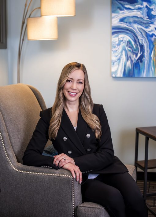 A woman with long, blonde hair is seated on a cushioned armchair, wearing a black blazer and black pants. She is smiling and has her hands resting on her lap. Behind her, there is abstract wall art and a modern floor lamp with three lights.