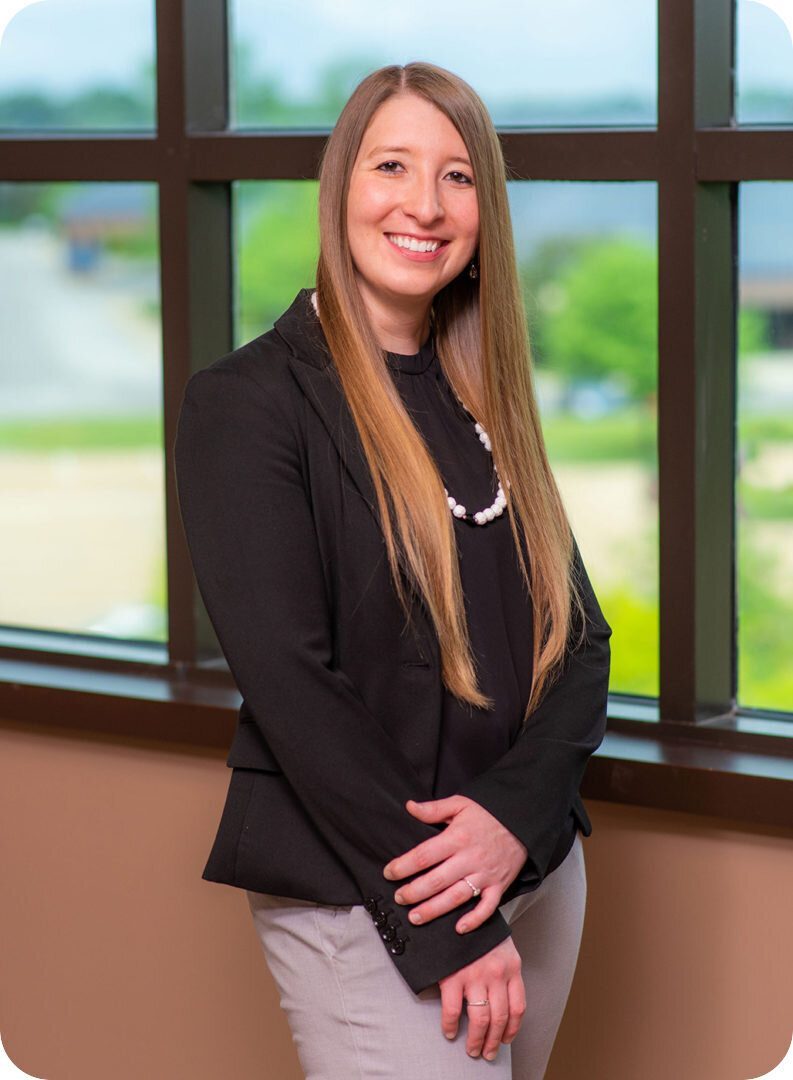 A woman with long, straight brown hair wearing a black blazer, black blouse, and light gray pants stands in front of a large window. She is smiling and has her hands clasped in front of her. The window shows a blurred outdoor background.